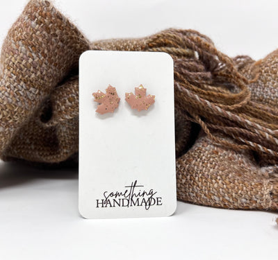 Pink and gold maple leaf stud earrings on a 'something handmade' card with a brown textured fabric background.